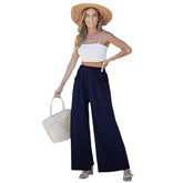 Woman wearing a white top and navy wide-leg pants with a straw hat and white bag on a white background