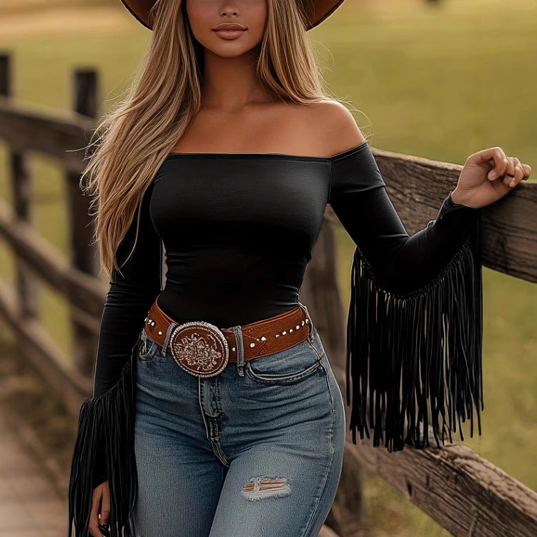 Woman wearing a black off-shoulder top with fringe sleeves and blue jeans, standing by a wooden fence.