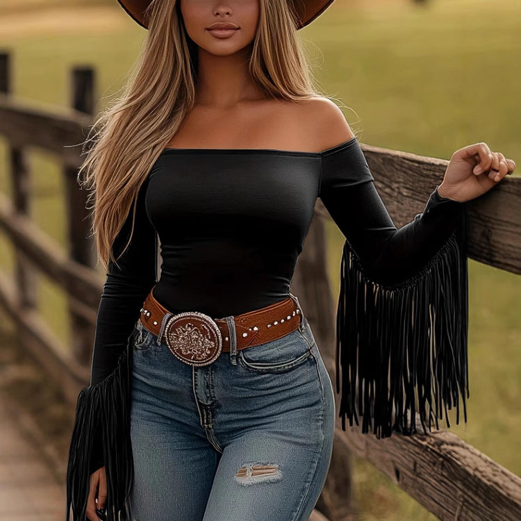 Woman wearing a black off-shoulder top with fringe sleeves and blue jeans, standing by a wooden fence.