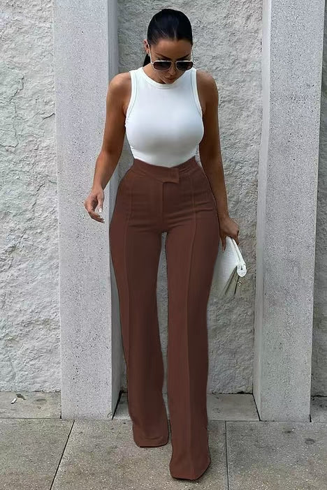 Woman wearing a white top and brown pants against a light stone wall.